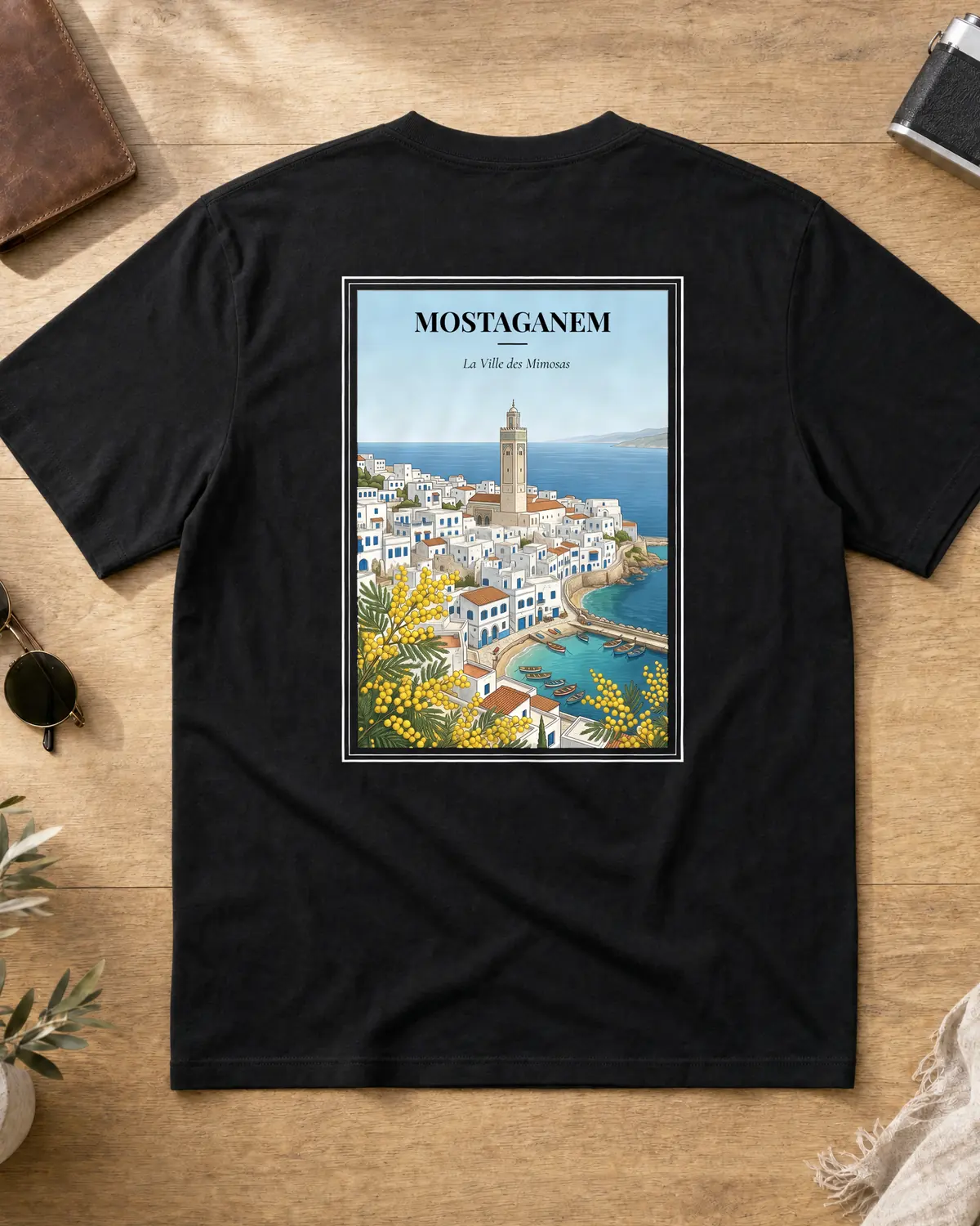 Black Mostaganem t-shirt back with vintage travel poster — whitewashed medina and red-tiled roofs tumbling to a turquoise cove, stone minaret at the crest, fishing boats in the harbour, yellow mimosa in the foreground under a calm blue sky