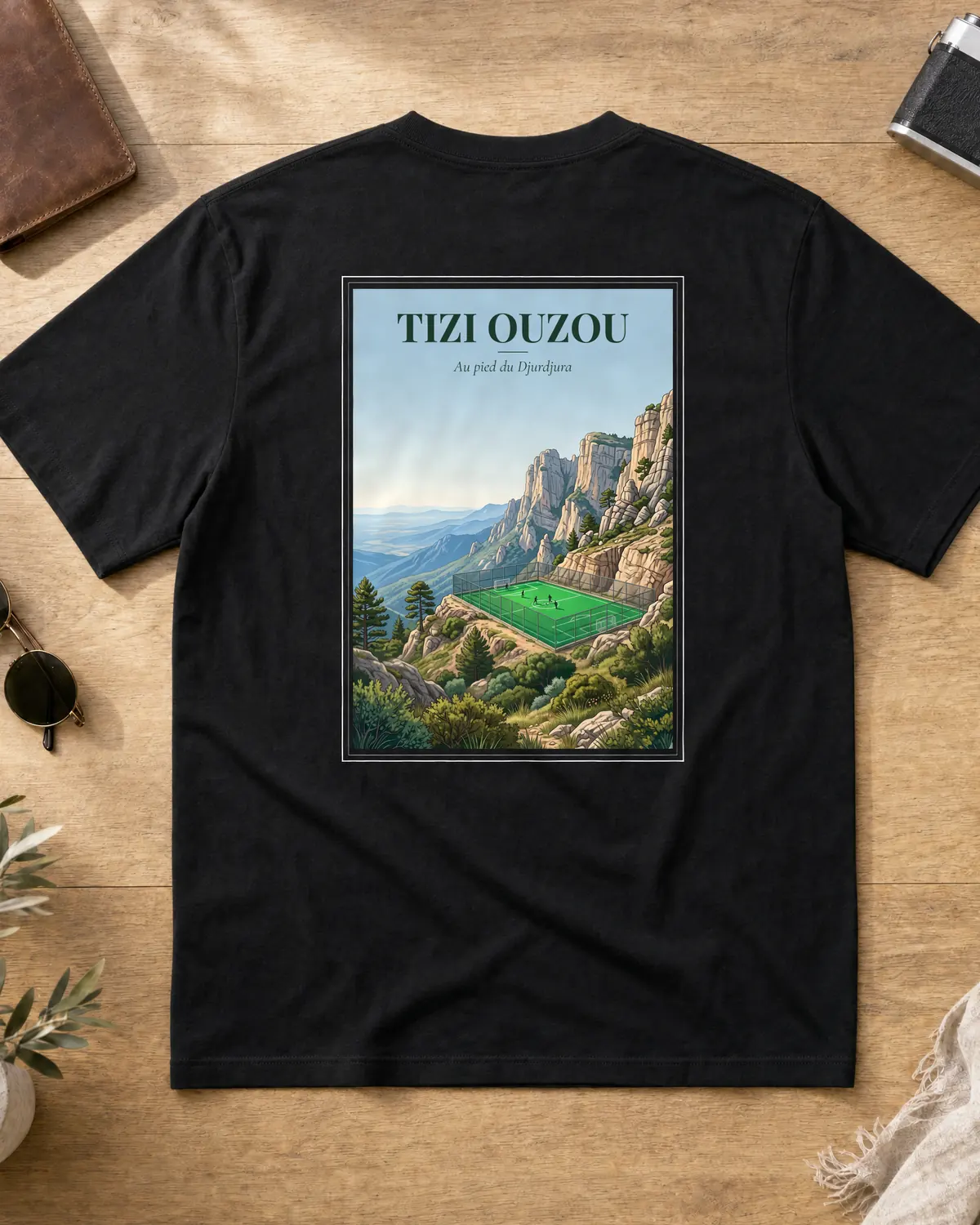 Black Tizi Ouzou t-shirt back with a vintage poster of the Djurdjura mountains, a football pitch in the valley, pine trees and the tagline Au pied du Djurdjura