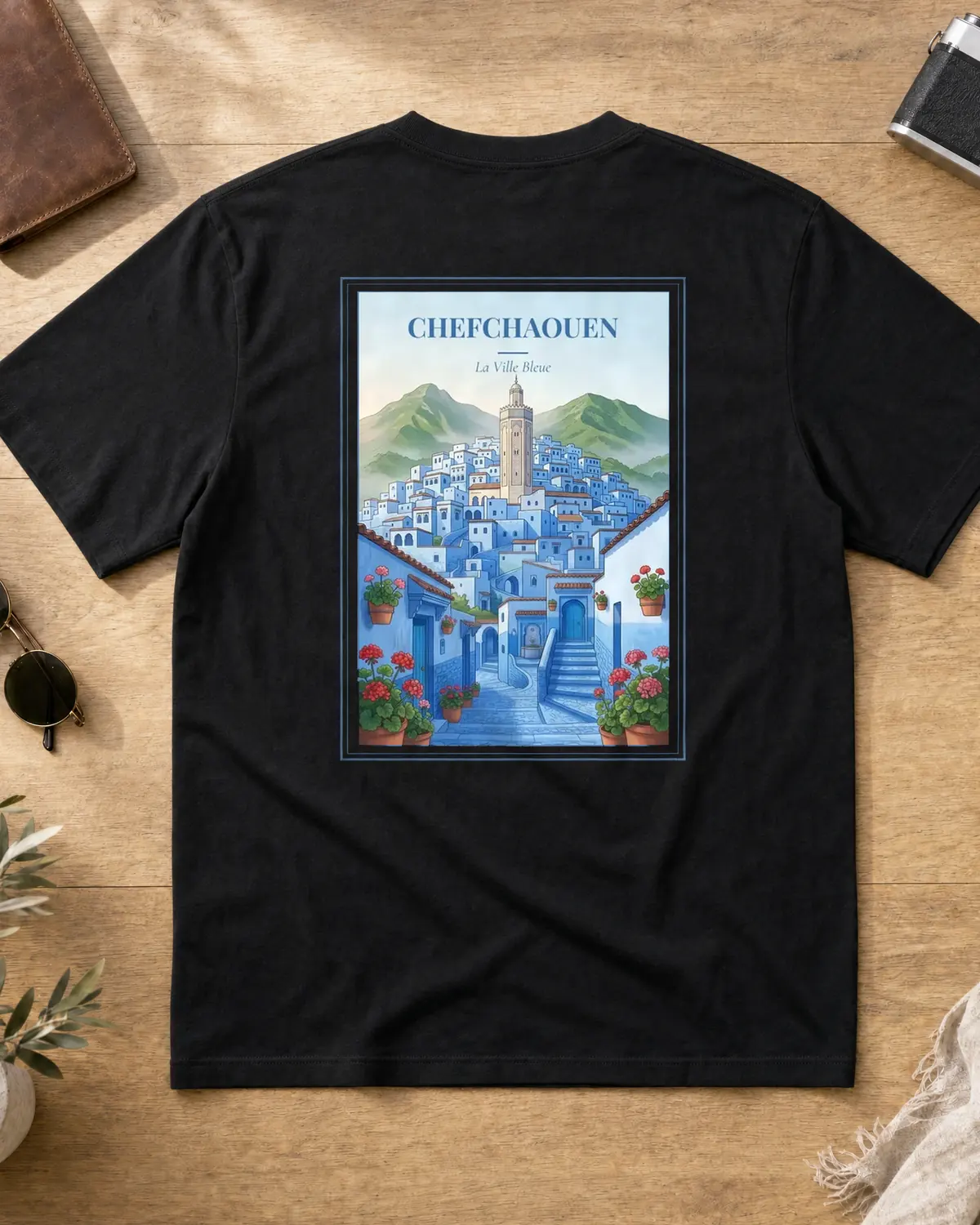 Black Chefchaouen t-shirt back with vintage travel poster — a blue-washed medina climbing the Rif hillside, a minaret and green mountains behind, and a stepped cobalt alley lined with terracotta pots of red geraniums